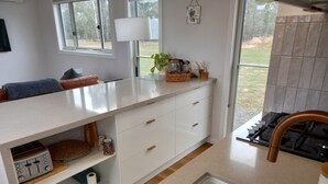 A modern kitchen with white cabinetry, a built-in gas stove, and a large window offering a view of the outdoors. - Lakeside Off-Grid Luxury Cabin Redman View (Lake Fyans)