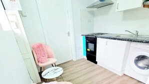 Cocina compacta con estufa, horno y lavadora. Hay una mesa redonda pequeña y una silla rosa con un plaid suave.