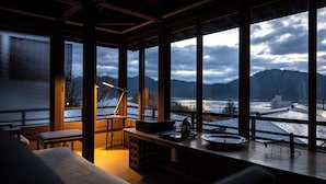 A room with a large window overlooking a snowy landscape and mountains. There is a desk with a lamp, a chair, and a small table with a teapot and cups. - Sekitei (Hatsukaichi)