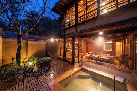 Un edificio tradicional japonés con una terraza de madera, un pequeño estanque y una zona de estar con cojines y una mesa baja.
