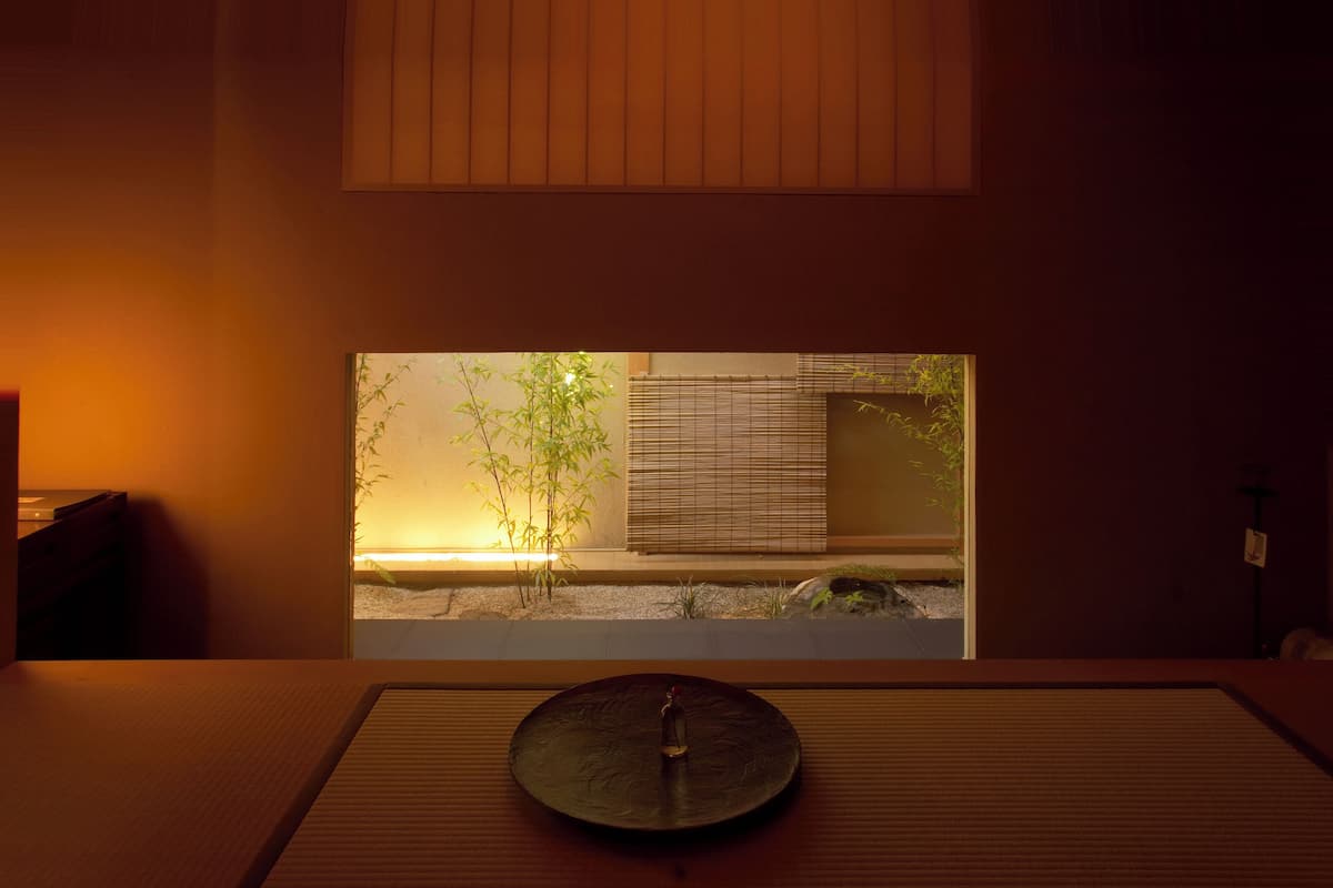 A traditional Japanese room with tatami flooring, a low table with a decorative item, and a view of a bamboo garden through a window.