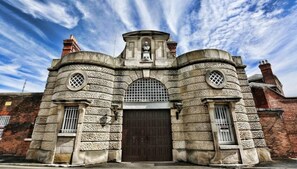 Ein historisches Steinhaus mit einem zentralen Rundbogenportal, das von zwei Rundfenstern mit dekorativen Gitterrosten flankiert wird, und einer Statue über dem Eingang.