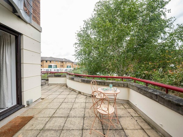 A small outdoor patio with a table and chair, a red railing, and a view of a building in the background.