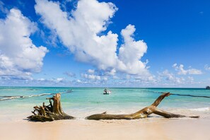 A beach with clear turquoise water, a boat in the distance, and a large tree trunk on the sand. - Rosa Hermosa Penthouse | Pool View • Near Bávaro (Punta Cana)