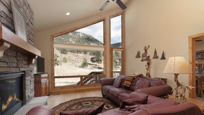 Una sala de estar con chimenea de piedra, un sofá de cuero, un ventanal con vistas a la montaña y un ventilador de techo de madera.