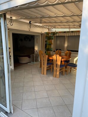 Un patio cubierto con una mesa redonda de madera y sillas, piso de baldosas y un cerramiento de vidrio corredero que da a un espacio interior.