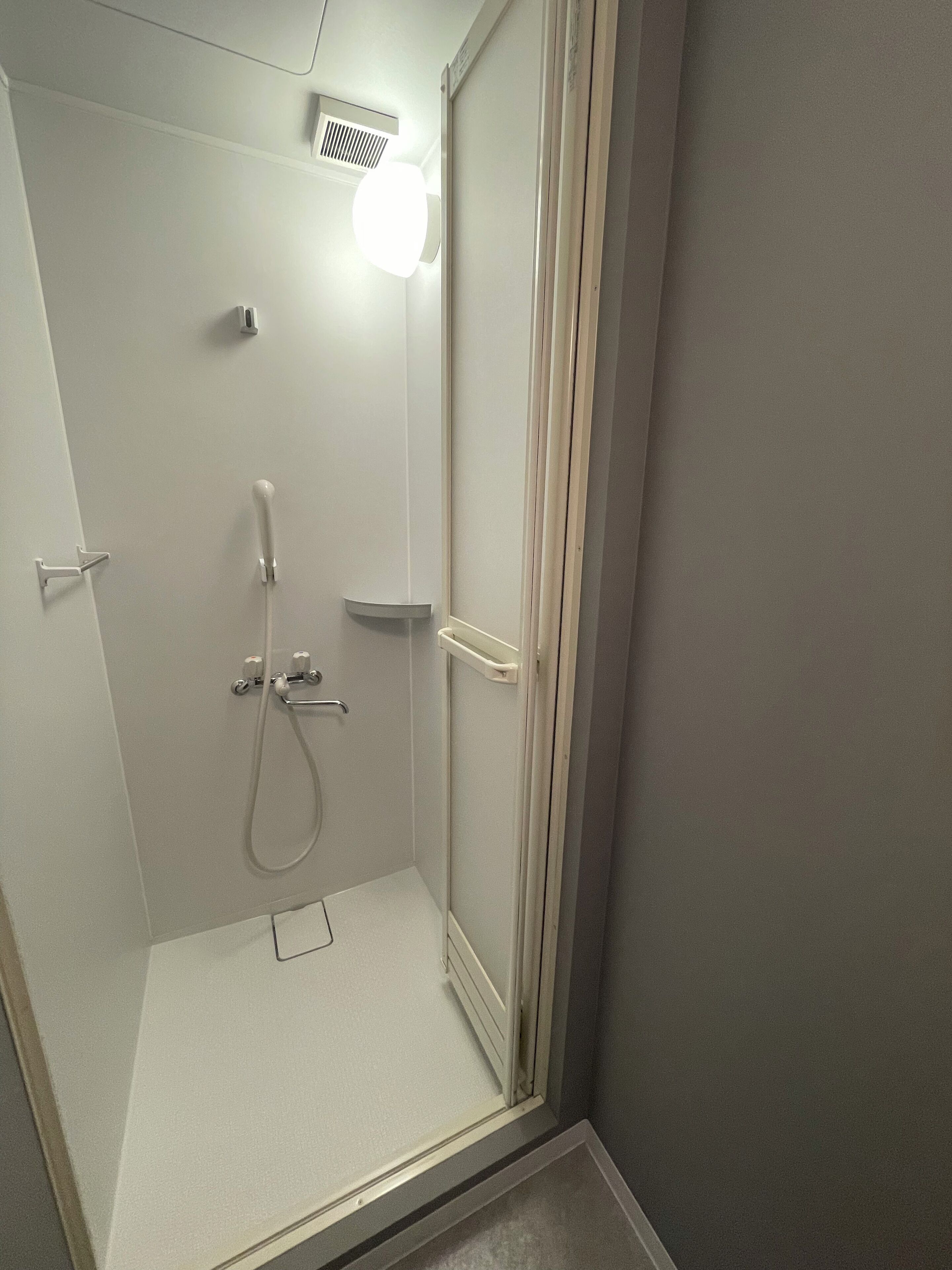 Un espacio de ducha pequeño y cerrado, con un cabezal de ducha manual, un calentador de pared y una lámpara sobre el calentador.
