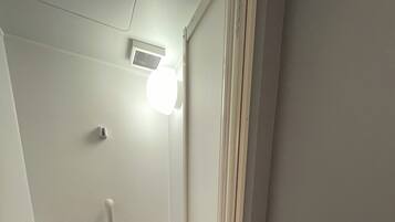 Un espacio de ducha pequeño y cerrado, con un cabezal de ducha manual, un calentador de pared y una lámpara sobre el calentador.