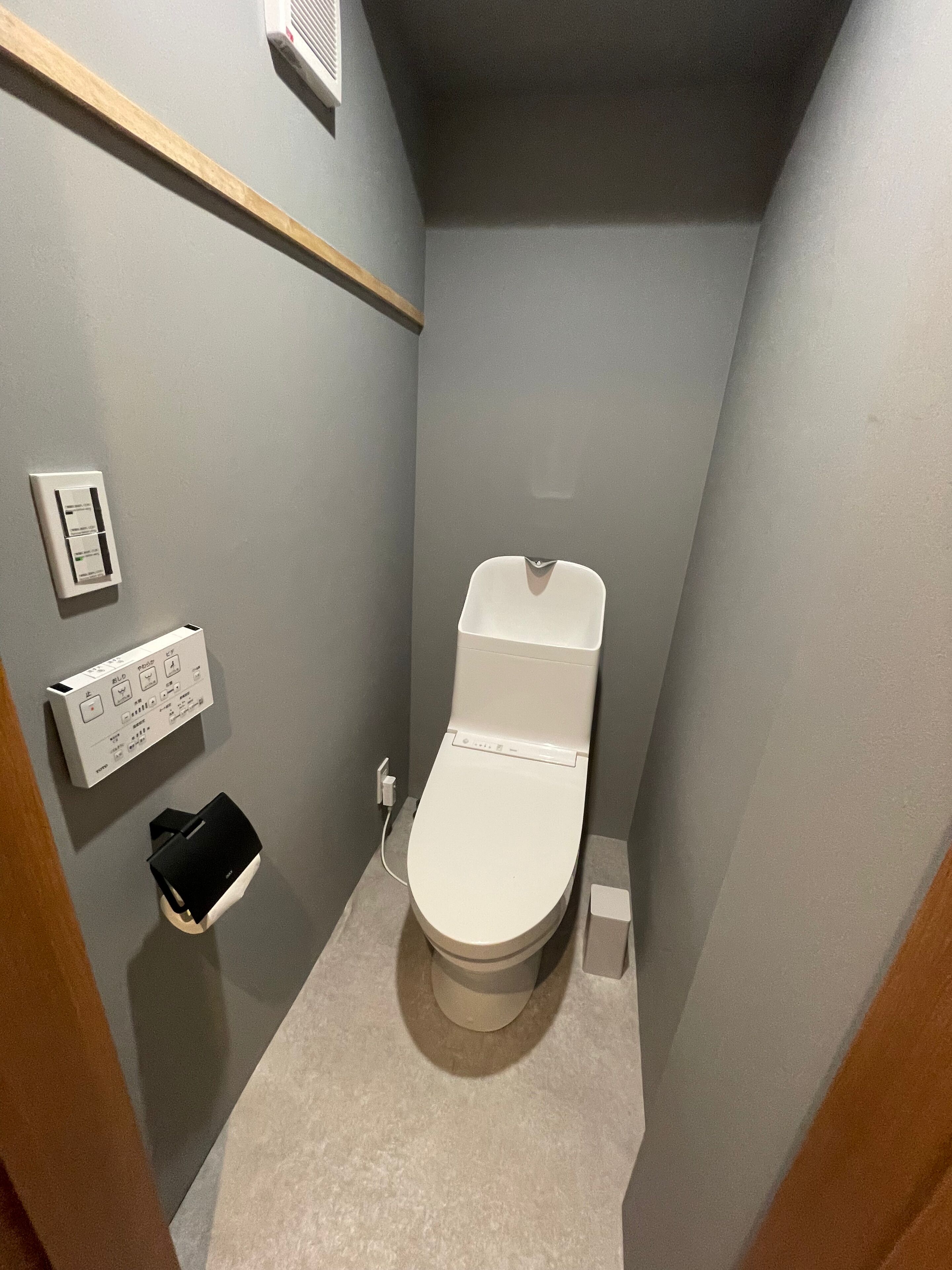 Een kleine badkamer met toilet, een inbouwspoelknop en een klein plankje met een zwart voorwerp.