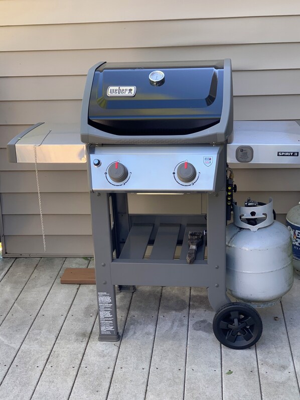 Una parrilla de gas Weber Spirit II con dos perillas de control, una cadena para colgar utensilios y un tanque de propano cerca.