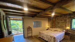 A wooden room with a bed, a wooden desk, a window with a view, and a stone wall. - El Pedrosu (Infiesto)