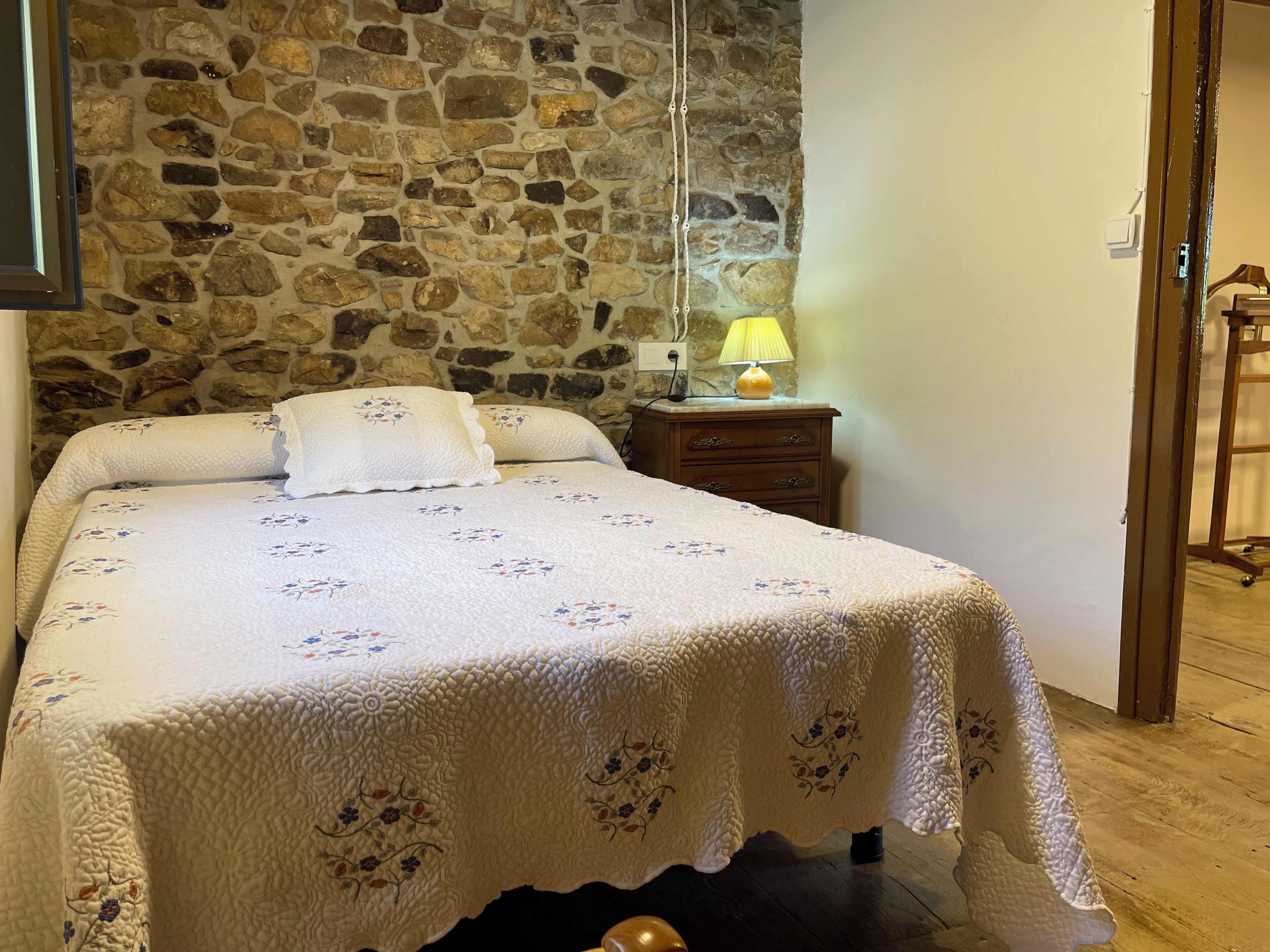 A bed with a floral bedspread, a stone wall, a nightstand with a lamp, and a wooden floor.