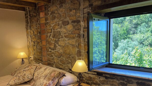 A bedroom with a stone wall, a bed with floral bedding, a nightstand with a lamp, and a window with a view of greenery. - El Pedrosu (Infiesto)