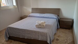 Een net opgemaakt bed met een bloemenbedekker en twee opgerolde handdoeken, een houten hoofdbord, een nachtkastje met een lade, en een betegelde vloer.