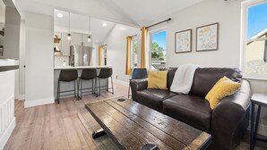 Un salon moderne avec un canapé en cuir marron, une table basse en bois et un coin cuisine doté d’appareils électroménagers en acier inoxydable et de tabourets de bar.