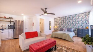 A compact living space with a kitchenette, a bed with a patterned bedspread, a red bench, and a ceiling fan. - Facing Collins Garden Park, 2nd Floor Studio (San Antonio)