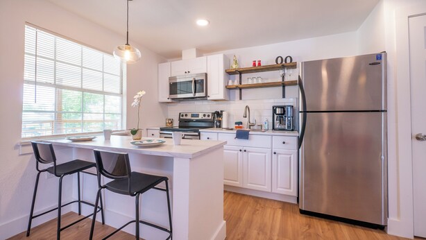 A modern kitchen with white cabinets, stainless steel appliances, a central island with bar stools, and a window with blinds. - Facing Collins Garden Park, 2nd Floor Studio (San Antonio)
