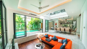 A modern living room with a large orange sofa, a coffee table, and a view of a swimming pool and greenery. - Villa Blue Water by GORO (Seminyak)