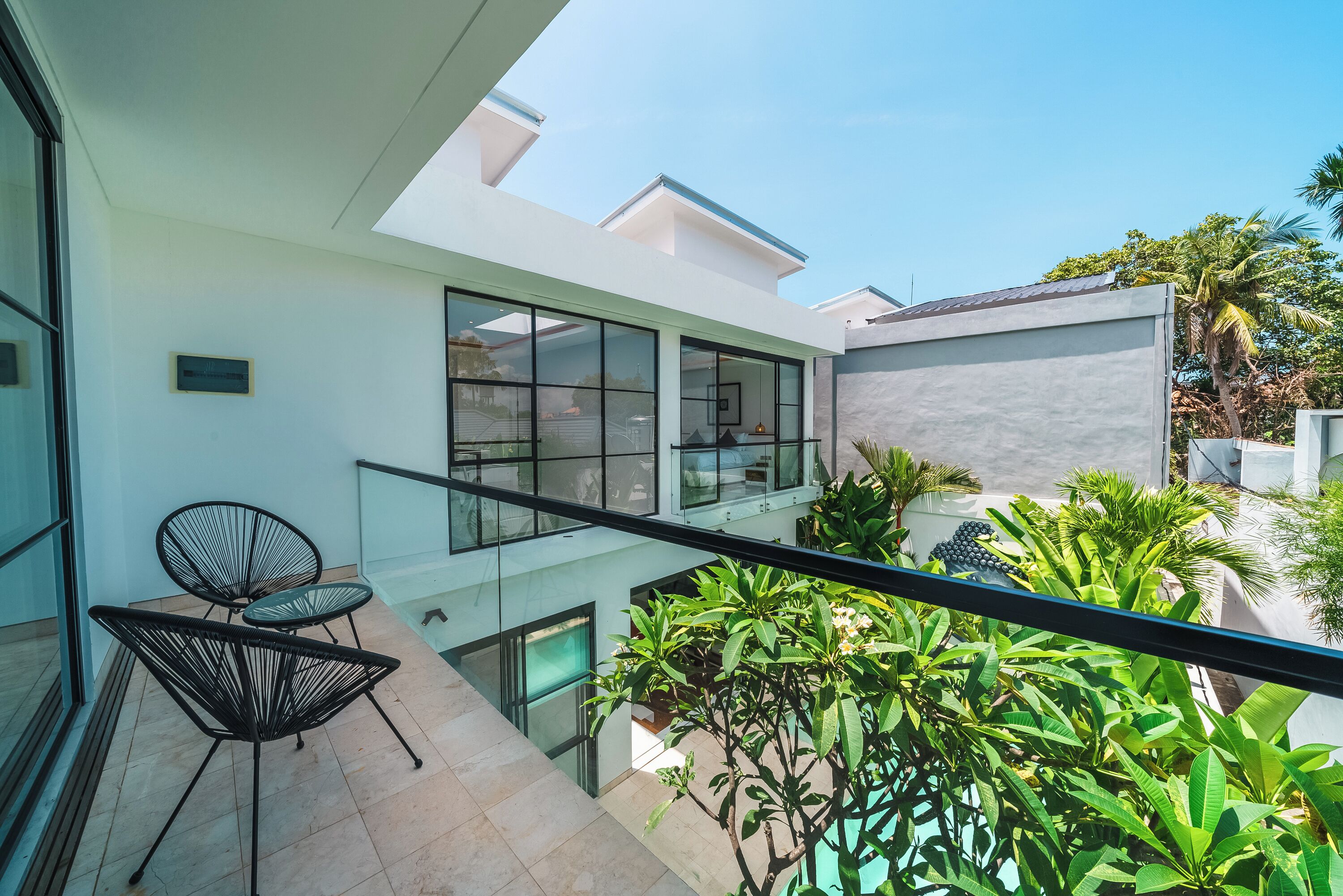 A modern balcony with glass railings, a seating area with chairs, and lush greenery.