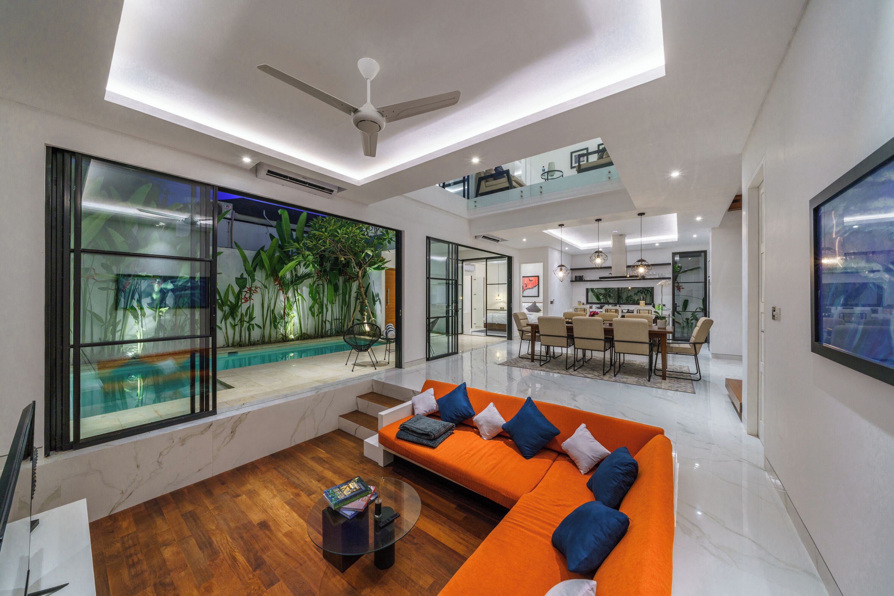 A spacious living area with a large orange sofa, a glass coffee table, and a view of a swimming pool and greenery.
