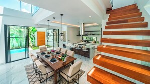A modern kitchen with a central island, a dining area with a wooden table and chairs, and a staircase with wooden steps. - Villa Blue Water by GORO (Seminyak)