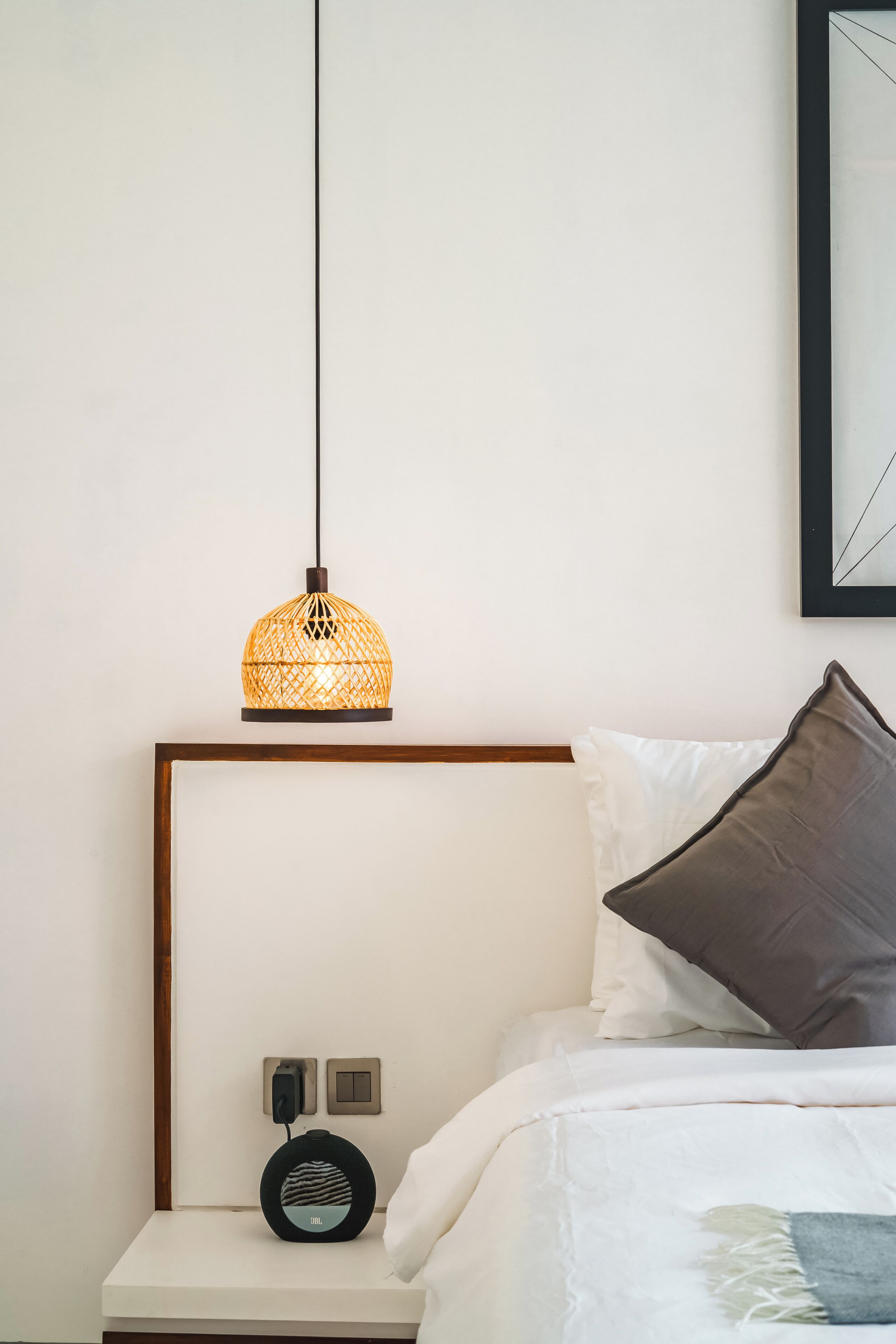 Un lit bien fait, avec un oreiller blanc et gris, une table de chevet sur laquelle se trouvent une lampe et un haut-parleur, et un tableau encadré accroché au mur.