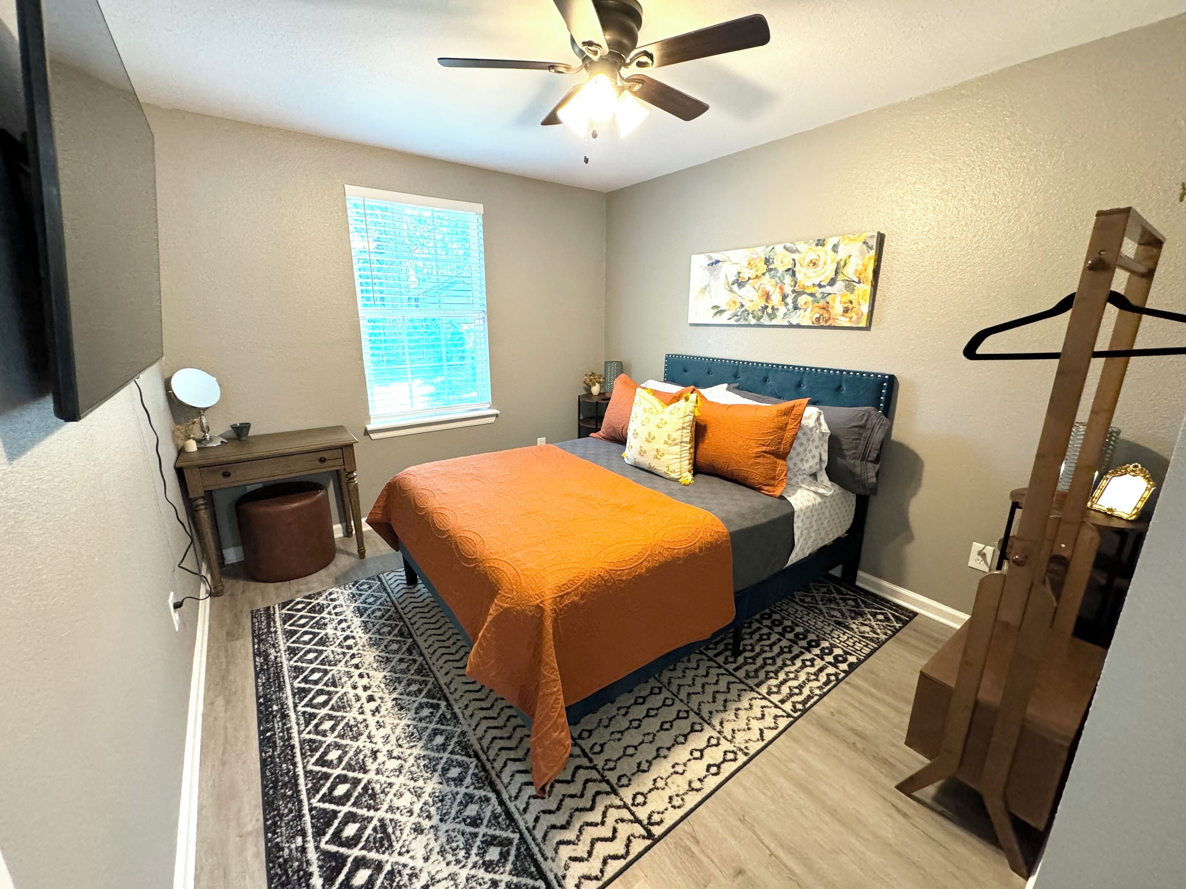 A bedroom with a bed, a bedside table, a mirror, a window, a ceiling fan, and a painting on the wall.