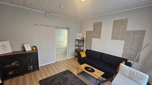 Een moderne woonkamer met een zwarte bank, een witte fauteuil, een houten salontafel en een donker vloerkleed. Er is een glazen deur die leidt naar een andere kamer, een klein plankje met decoratieve objecten, en een wand met geometrische patronen.