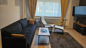Een moderne woonkamer met een zwarte bank, een houten salontafel, een televisie en een raam met gordijnen.