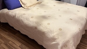 Un lit bien fait, avec une couvre-lit à motifs et des oreillers, une tête de lit capitonnée, une table de chevet blanche avec une lampe, et une fenêtre ornée de rideaux légers.