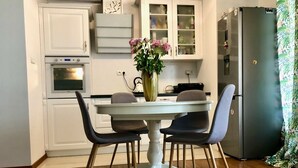 Un coin repas avec une table blanche et quatre chaises, un vase avec des fleurs, et une cuisine avec des meubles blancs et un réfrigérateur.