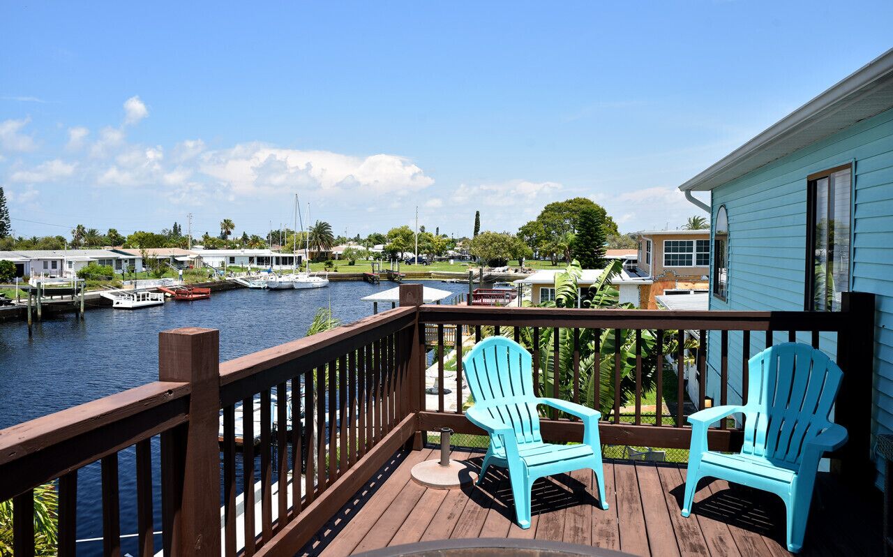 Um deck de madeira com duas cadeiras Adirondack turquesa de frente para um calçadão com barcos e um céu azul límpido.