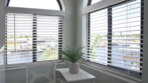 Une pièce d’angle avec un bureau blanc, une plante en pot et une vue sur un parking, visible à travers de grandes fenêtres équipées de stores.