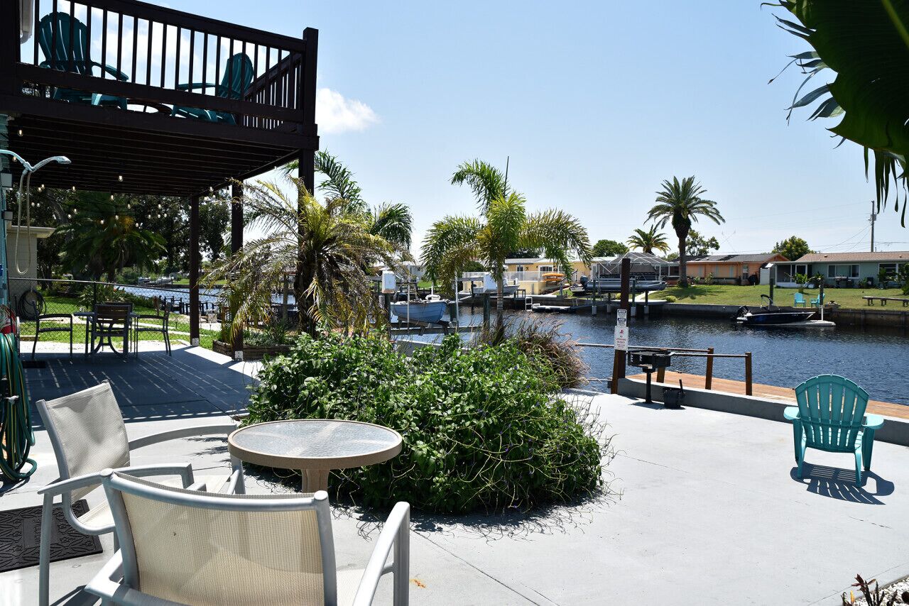 An outdoor patio with chairs and a table, overlooking a body of water with boats and houses in the background.