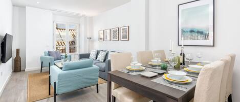 Una sala de estar moderna con comedor, que incluye un sofá, una mesa de comedor para cuatro personas y un televisor de pantalla plana montado en la pared.