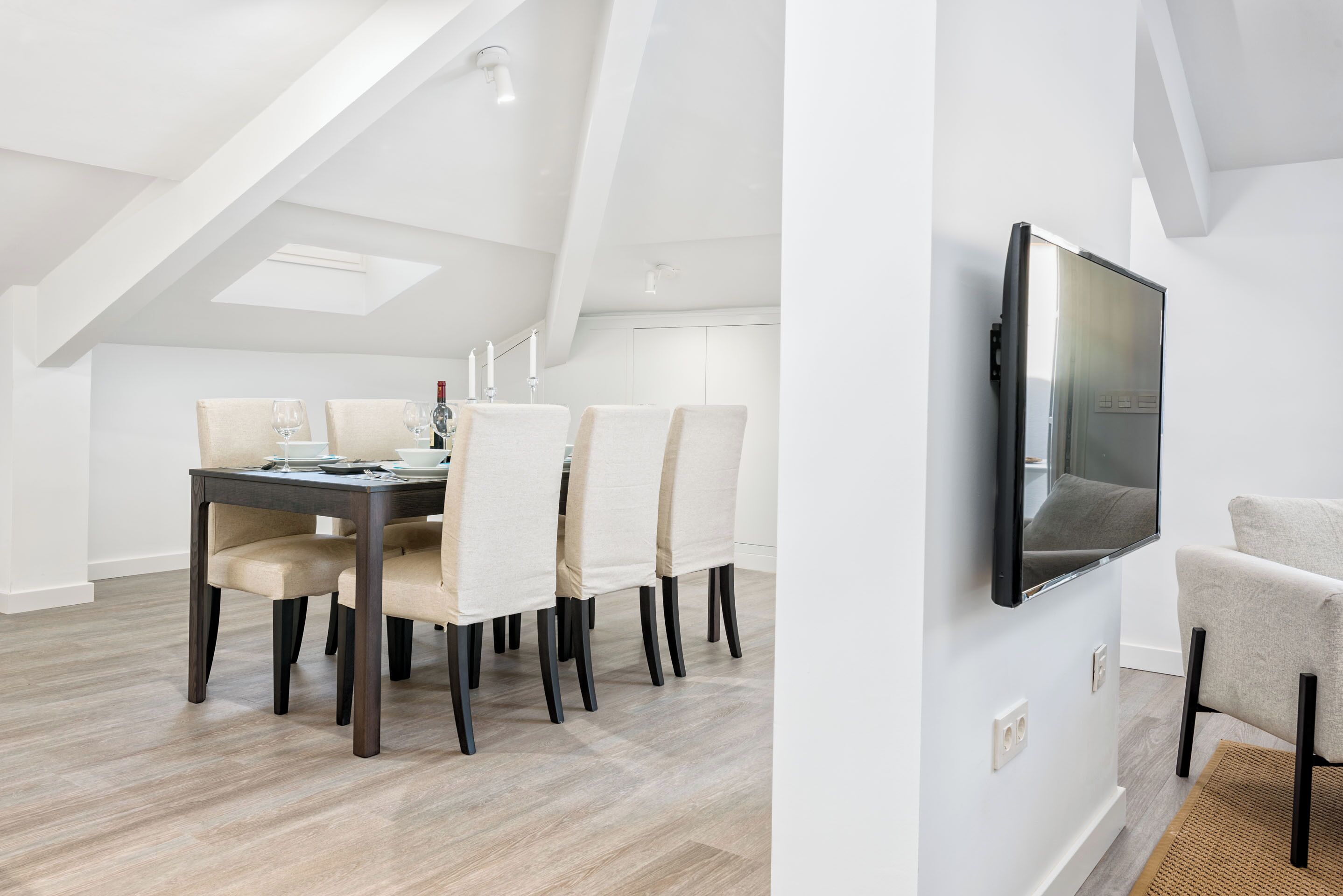 A modern dining area with a table and chairs, a flat-screen TV mounted on the wall, and a sloped ceiling with skylights.