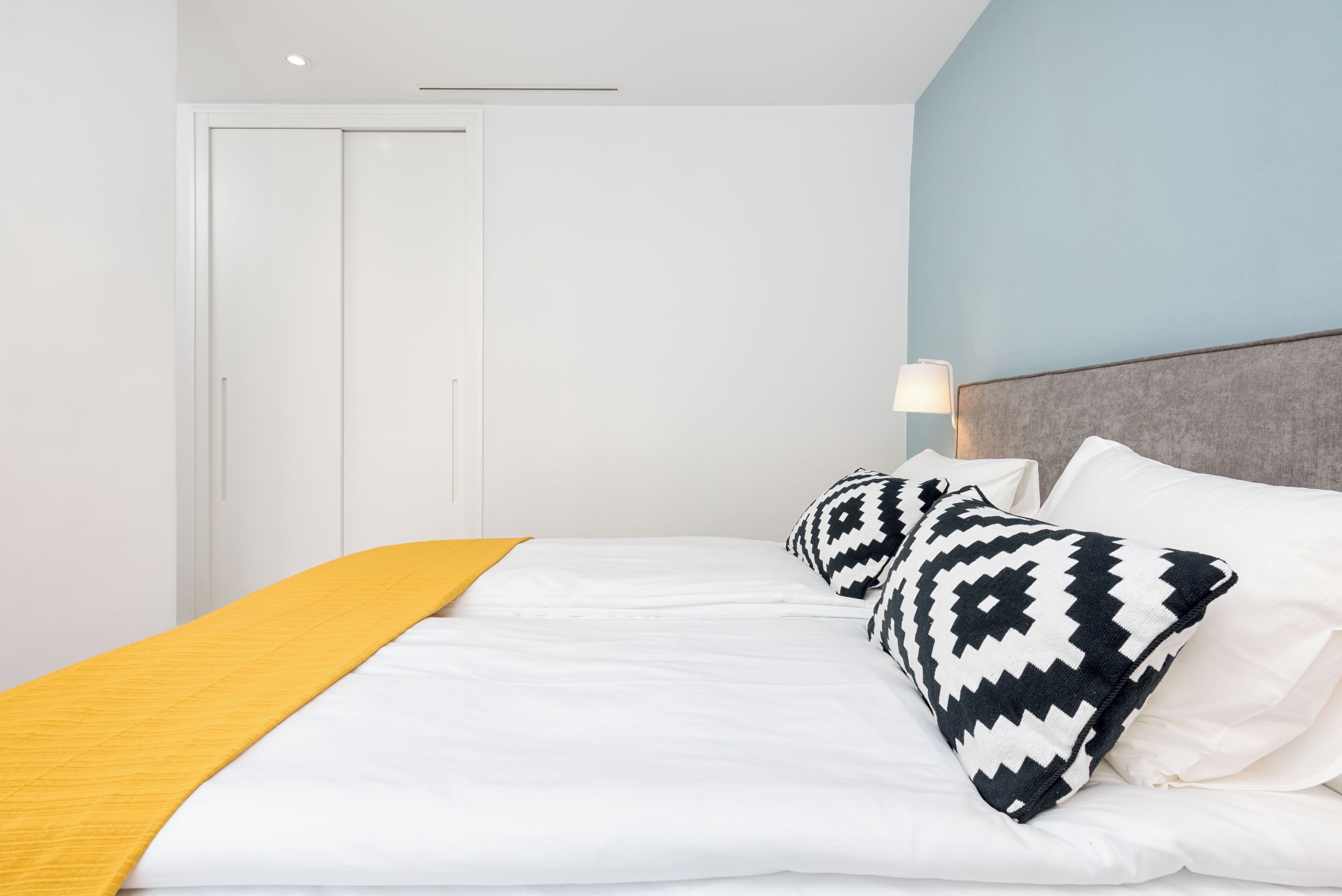 Una cama bien hecha con ropa de cama blanca y una manta amarilla, un cojín estampado y una lámpara de noche en una mesita.