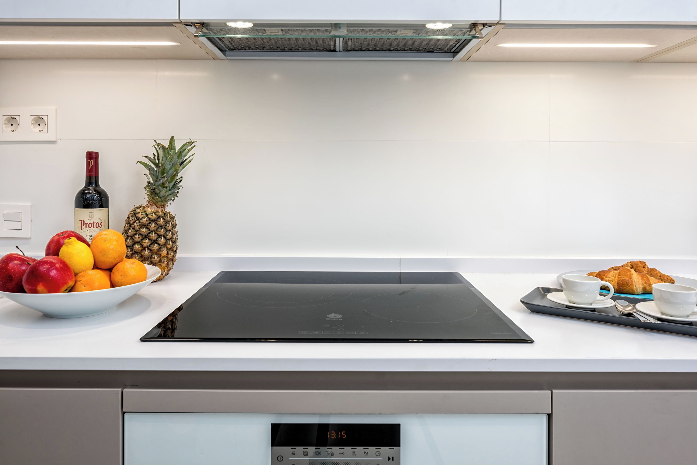 A modern kitchen with a black induction cooktop, a bowl of fruits, a bottle of wine, and a tray with croissants and cups.