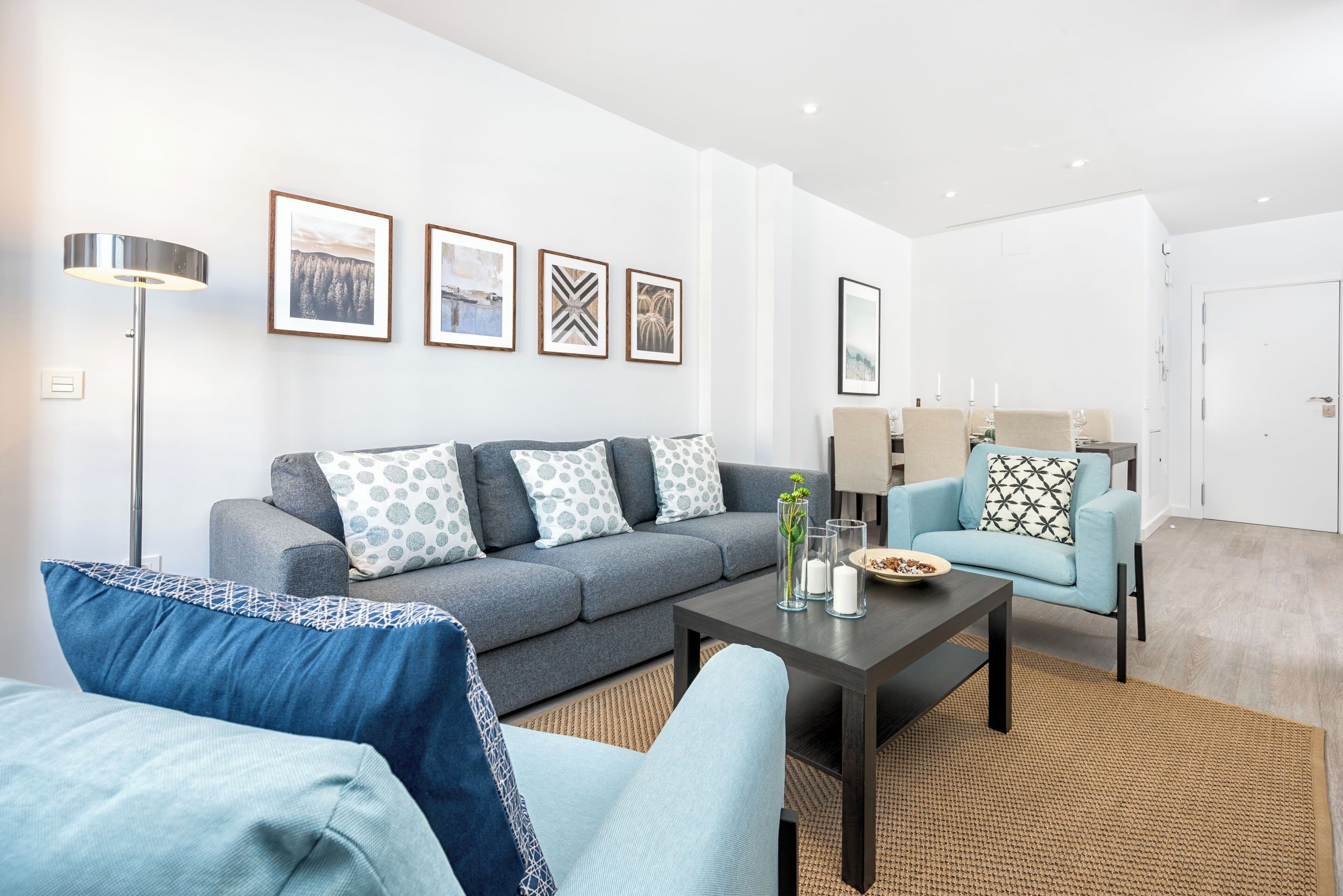 A modern living room with a grey sofa, blue armchairs, and a coffee table. There are framed pictures on the wall and a floor lamp.
