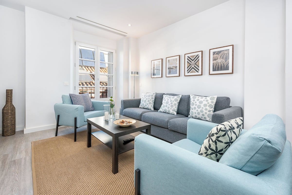 A modern living room with a grey sofa, blue armchairs, and a coffee table. There are framed pictures on the wall and a large window with a view of a building.