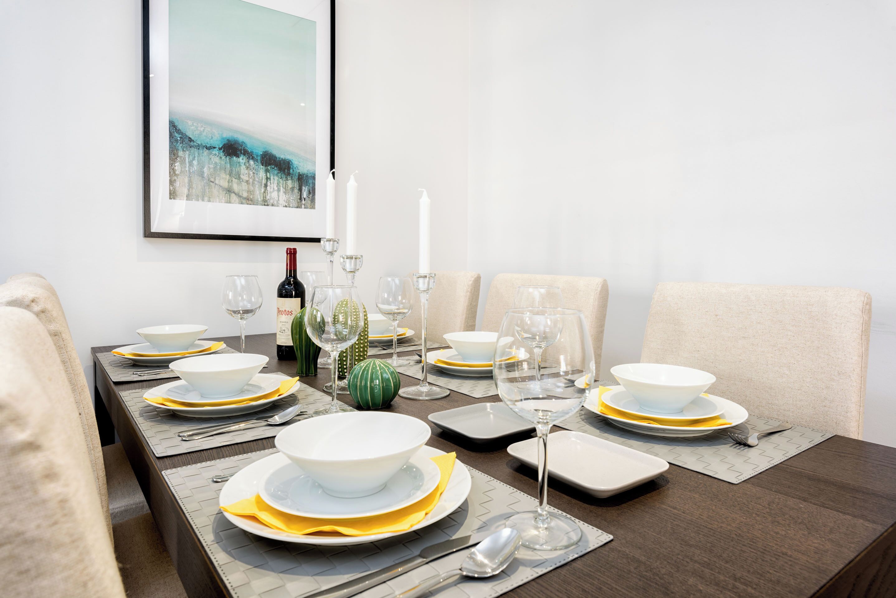 A dining table set with white plates, yellow napkins, and clear glassware, with a framed picture above and a bottle of wine on the table.