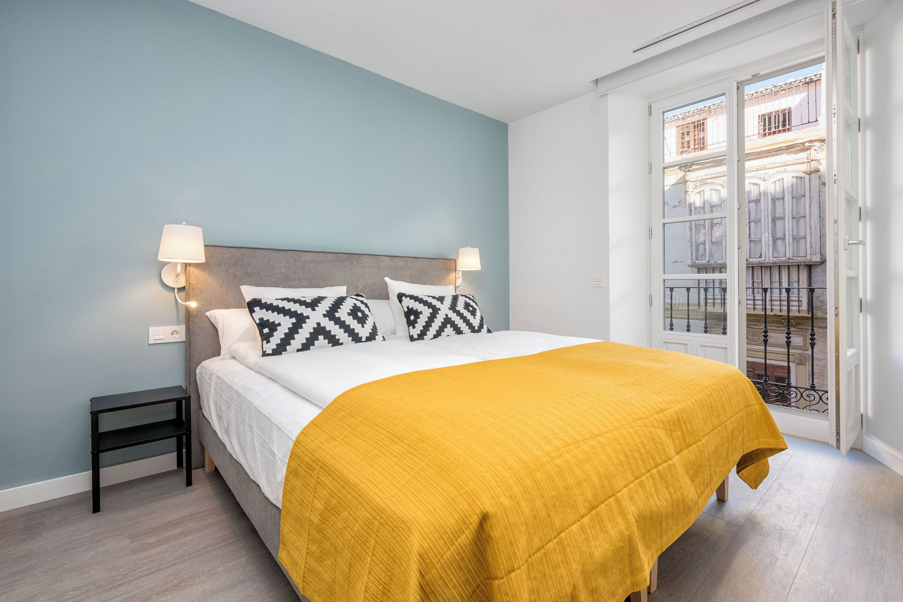 A bedroom with a bed, two bedside lamps, a nightstand, a yellow bedspread, and a view of a balcony with a railing.