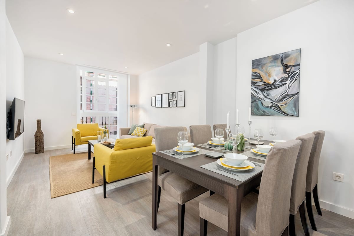 A modern dining area with a wooden table set for four, surrounded by upholstered chairs. A yellow sofa and a large window with blinds are visible in the background.