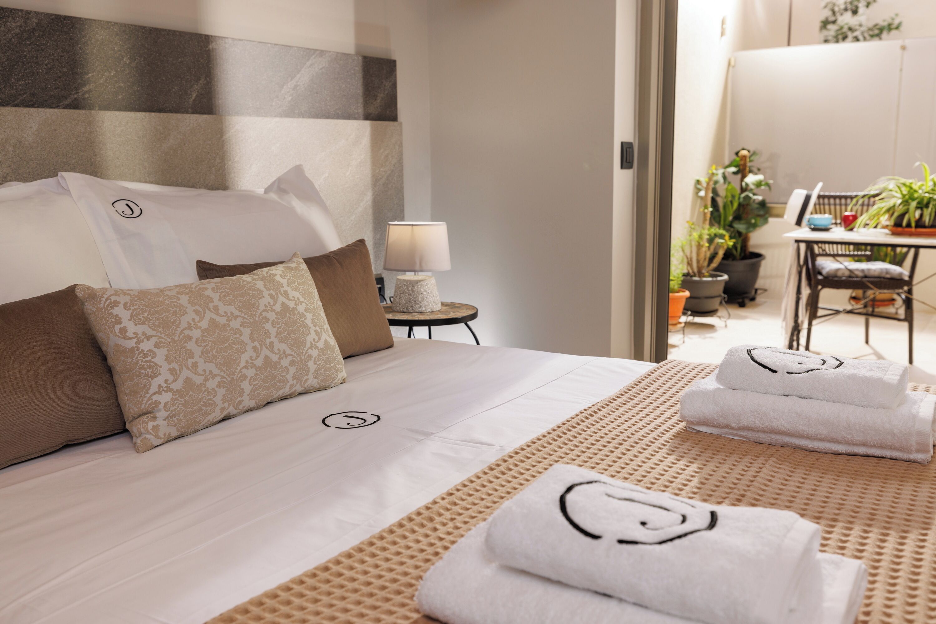 Ein Hotelzimmer mit Bett, Kissen, Nachttisch mit Lampe, einem kleinen Tisch mit Stuhl und Topfpflanzen.