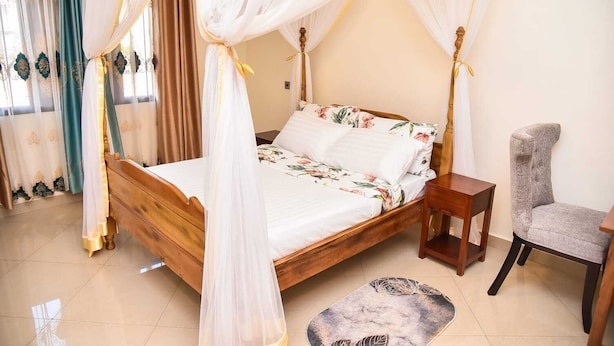 A four-poster bed with a floral bedspread, a wooden nightstand, a grey upholstered chair, and a patterned rug. - KCube Villas Kampala (Kampala)