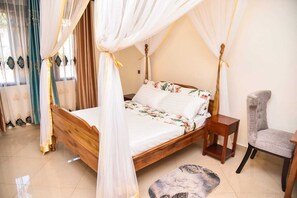 A four-poster bed with a floral bedspread, a wooden nightstand, a grey upholstered chair, and a patterned rug. - KCube Villas Kampala (Kampala)