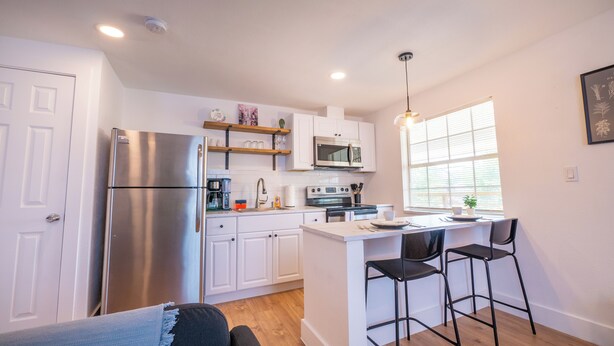 A compact kitchen with white cabinets, a stainless steel refrigerator, a microwave, and a coffee maker. There is a small island with bar stools and a window providing natural light.
