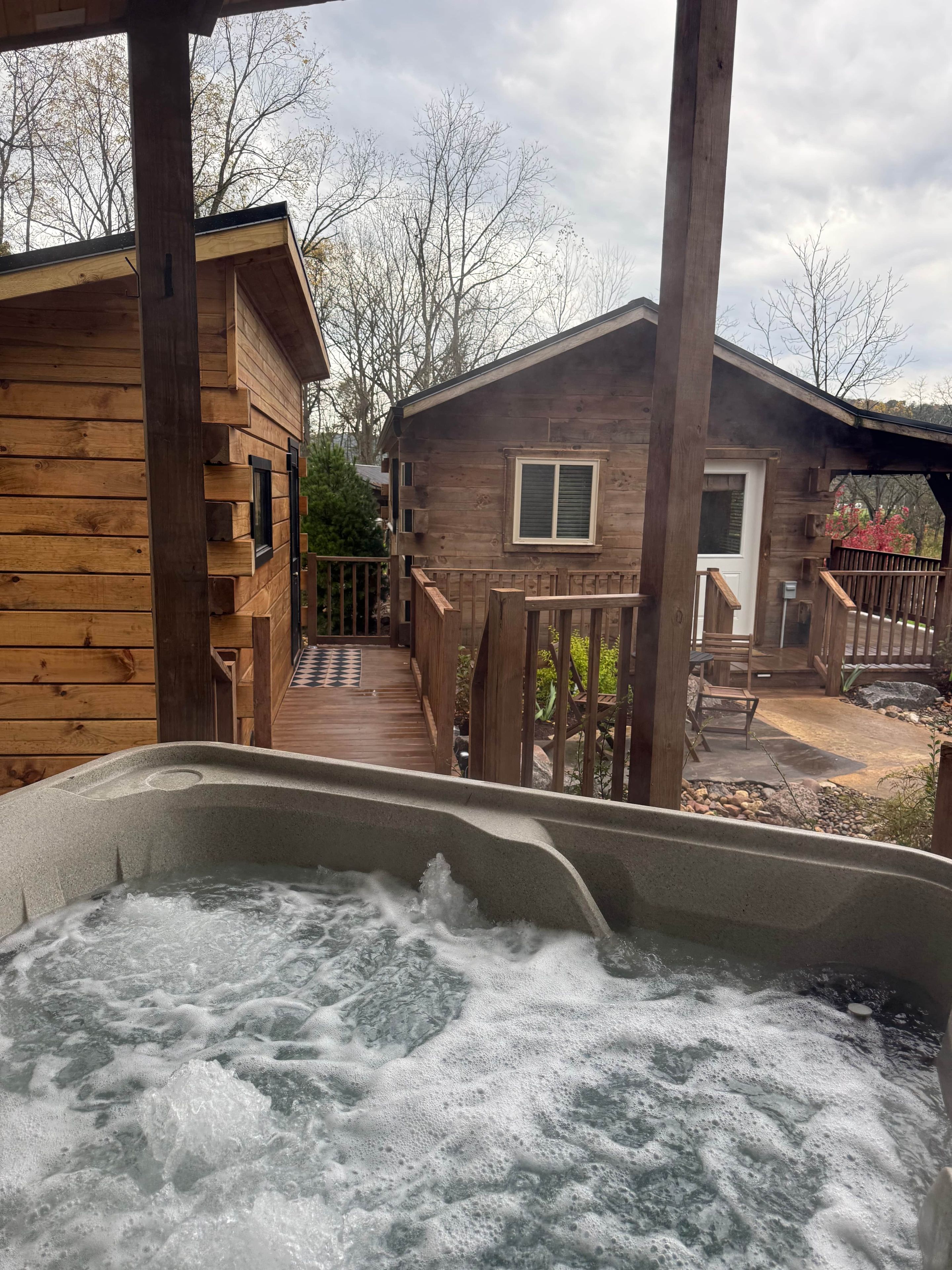 Una cabaña de madera con un jacuzzi en primer plano, una terraza y vistas a los árboles circundantes.