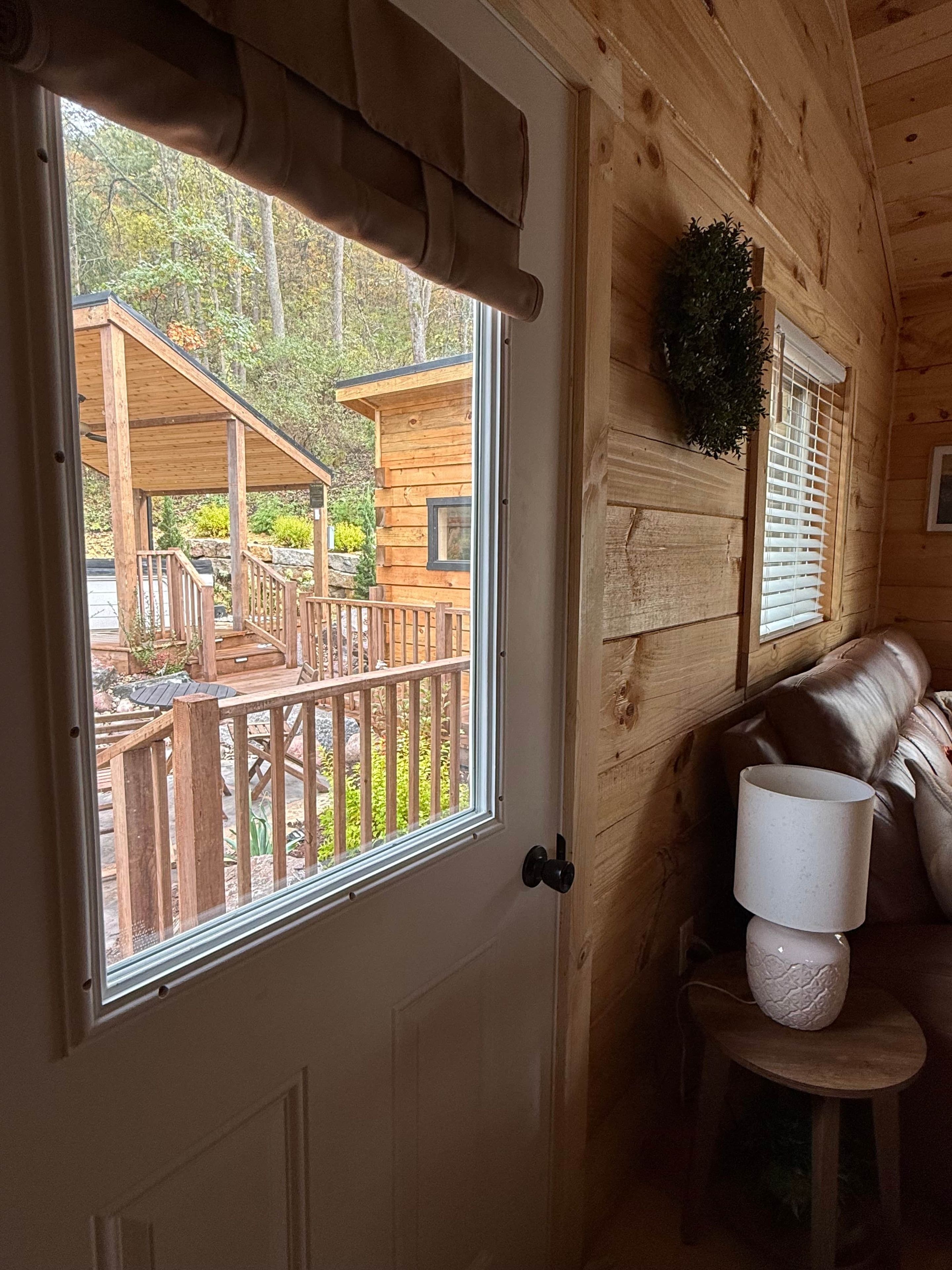L'intérieur d'un chalet en bois, avec une porte donnant sur une terrasse extérieure, un canapé en cuir et une petite table avec une lampe.
