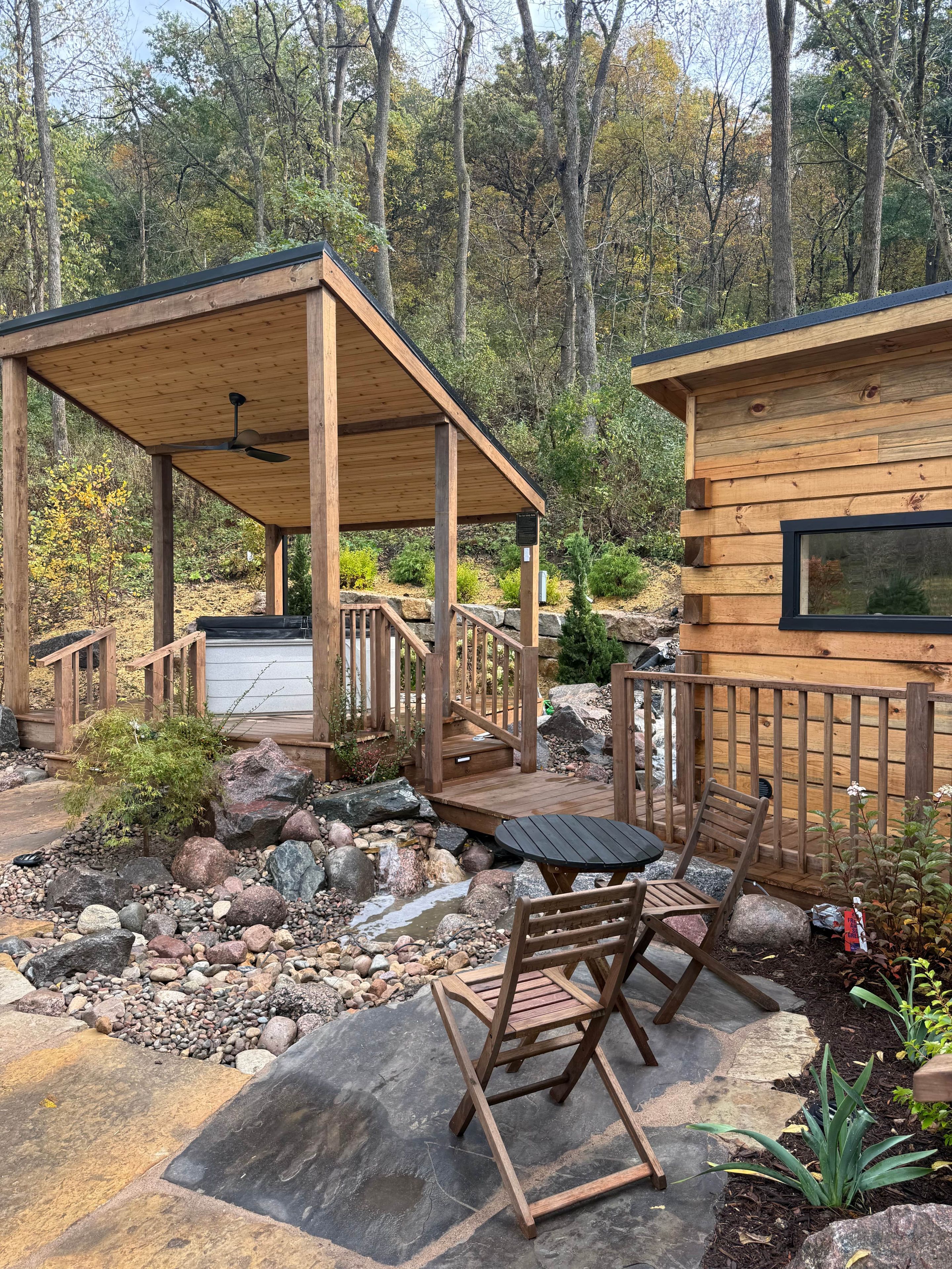 Een houten hut met een overdekt terras, buitenmeubels en uitzicht op het omliggende bos.